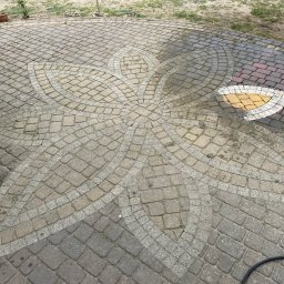 Krzysztof Kuliński - Czyszczona kostka brukowa z wzorem kwiatowym. Część kostki jest mokra, ukazując efekt czyszczenia. Widoczny wąż i fragment samochodu.