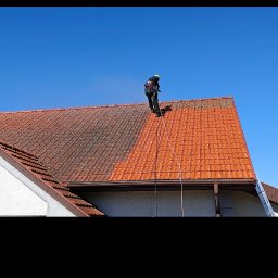 Krzysztof Kuliński - Częściowo wyczyszczony dach z czerwonej dachówki. Widoczna różnica między czystą a zabrudzoną powierzchnią. Pracownik w uprzęży na dachu.