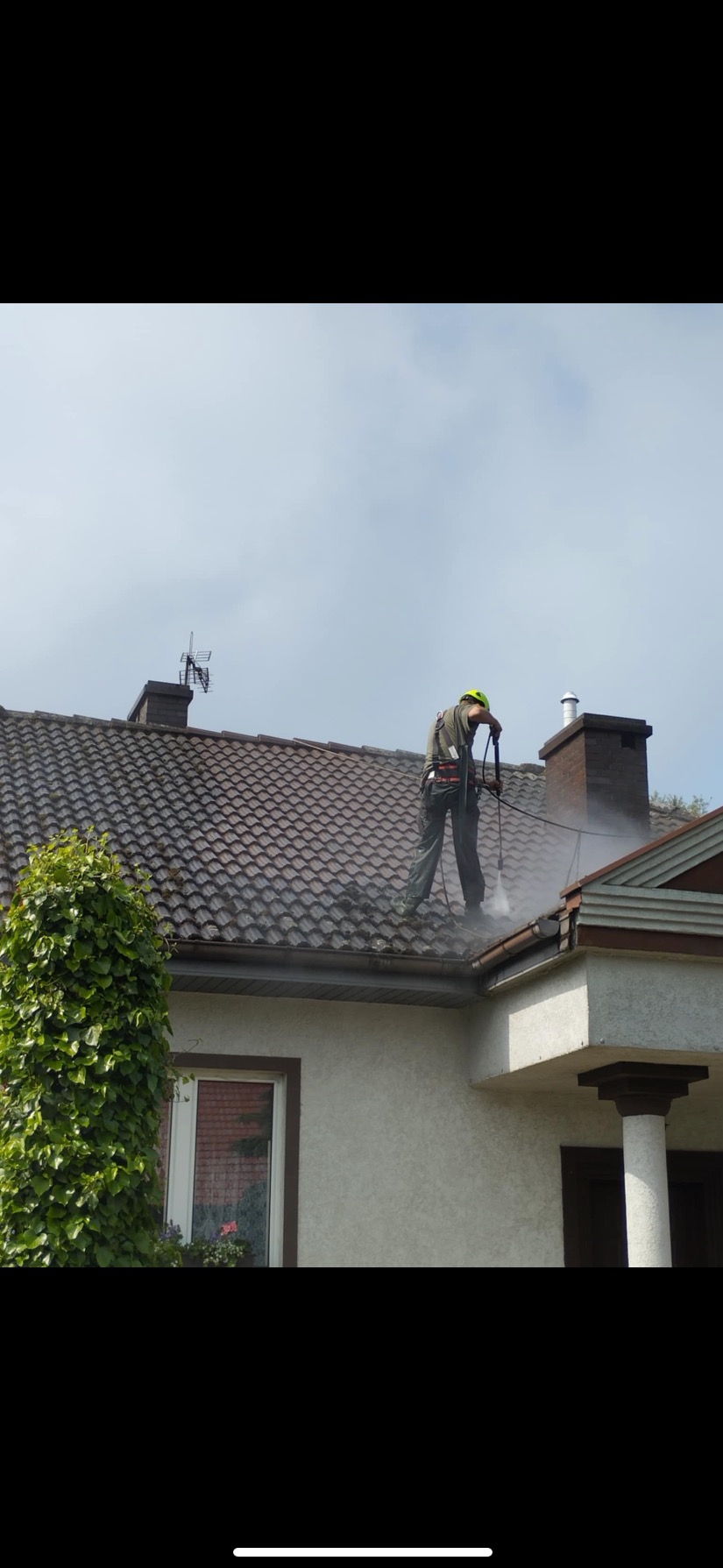 Pracownik w kasku myje dach z dachówki za pomocą myjki ciśnieniowej. Widoczne zacieki i strumień wody. Na dachu komin i antena.