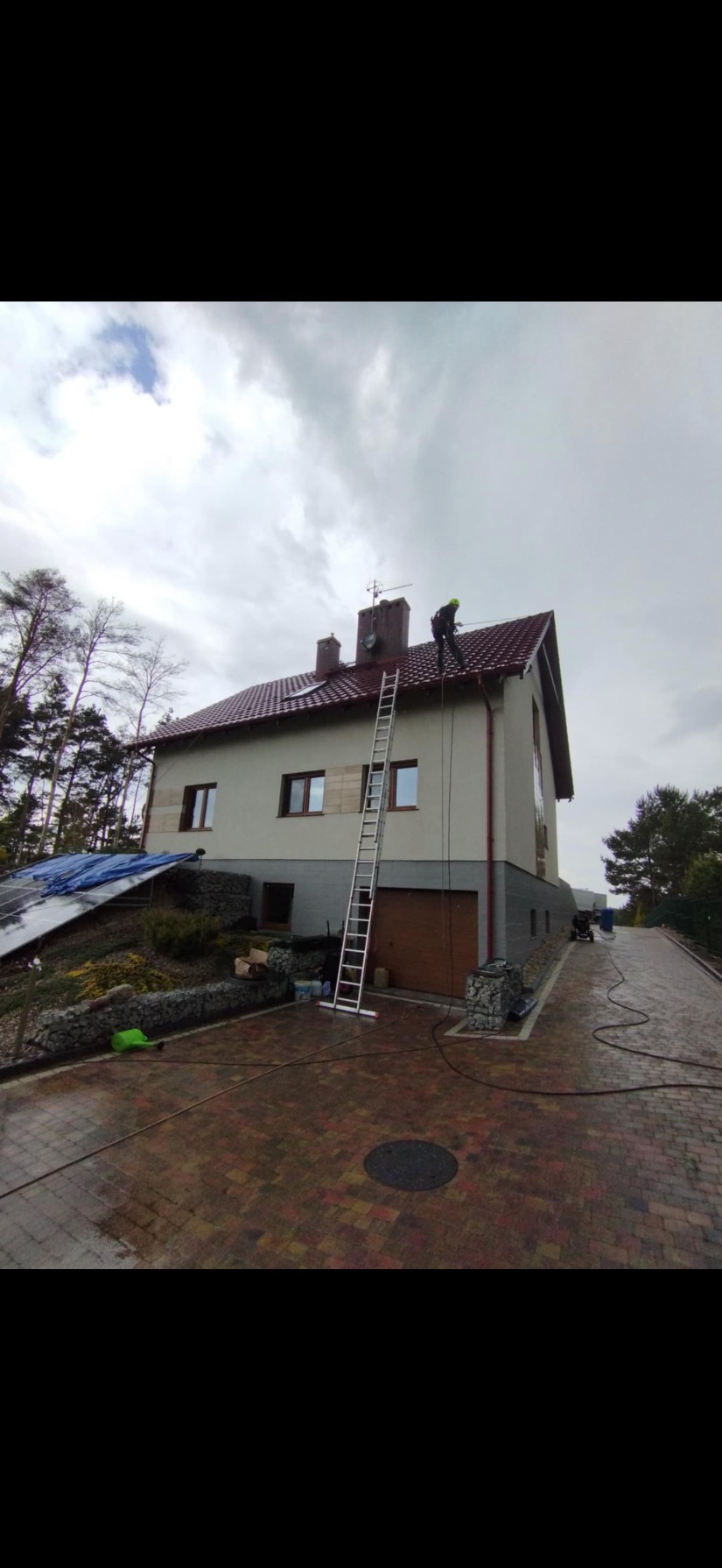 Pracownik na dachu budynku z czerwonym dachem, zabezpieczony linami, obok drabina. Widoczny mokry bruk i otoczenie domu. Prace na wysokościach.