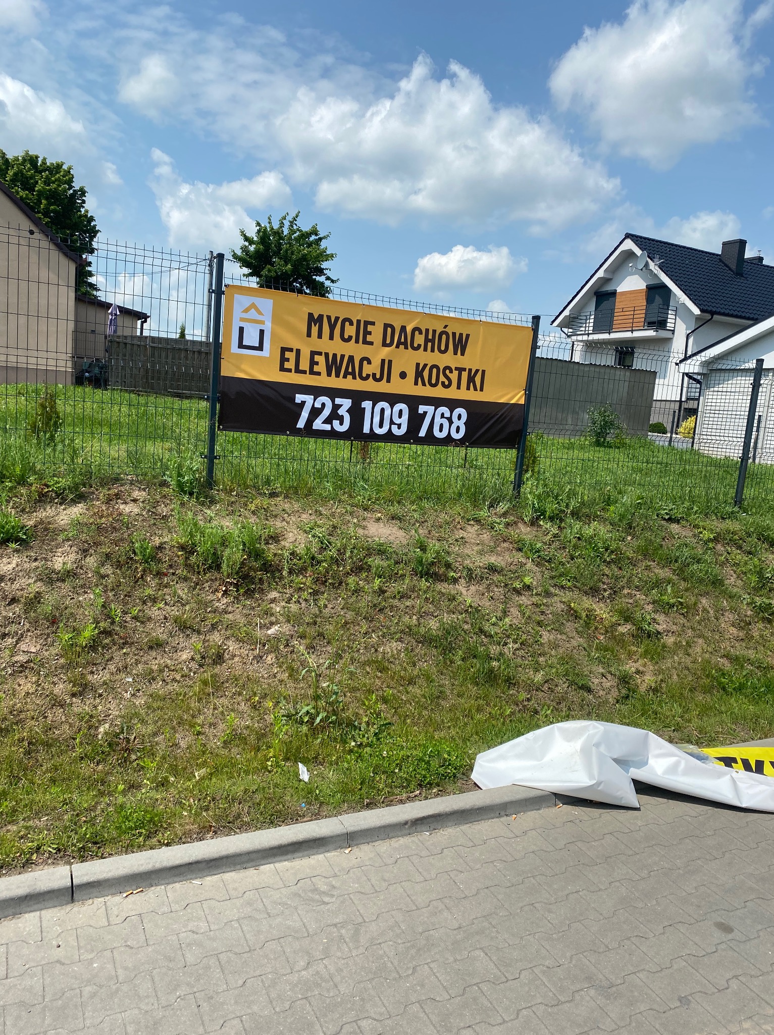 Żółto-czarny baner reklamowy z numerem telefonu i napisem 'Mycie dachów, elewacji i kostki' na tle ogrodzenia, trawnika i domów w słoneczny dzień.