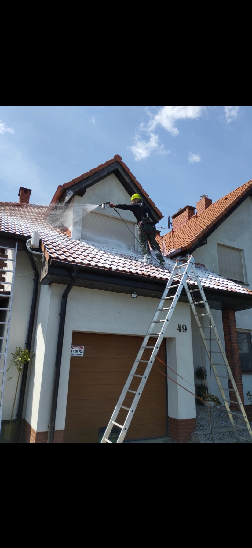 Pracownik w kasku, na dachu domu, myje go pod ciśnieniem. Widoczne drabiny i fragment elewacji. Słoneczny dzień.