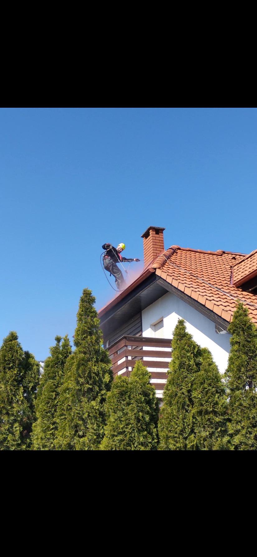 Pracownik w kasku i uprzęży czyści dach z czerwonej dachówki za pomocą myjki ciśnieniowej. W tle błękitne niebo i zielone tuje.