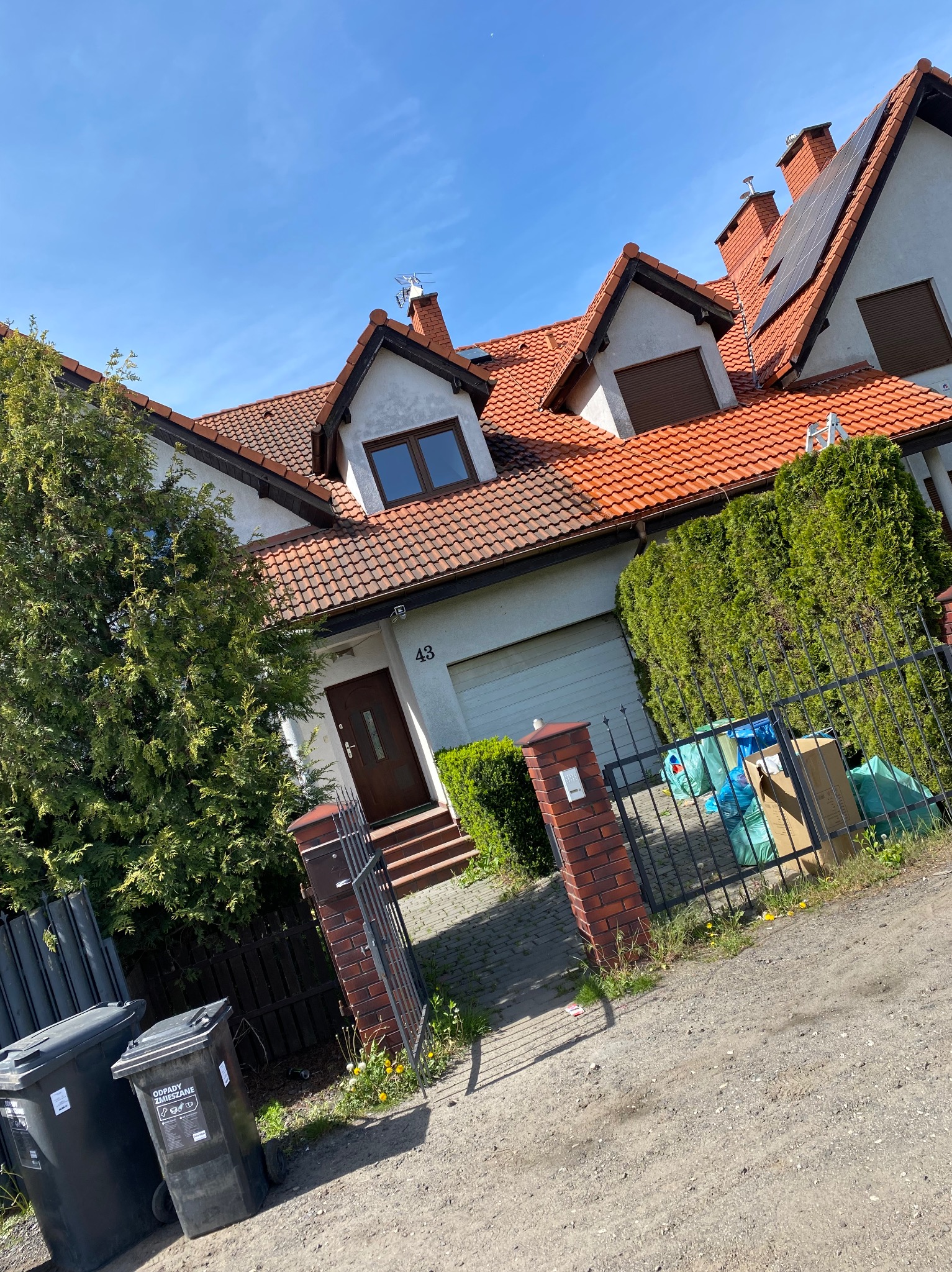 Dom jednorodzinny z czerwoną dachówką, panele słoneczne, zadbany ogród i śmietniki przed posesją. Ujęcie z dołu, w słoneczny dzień.