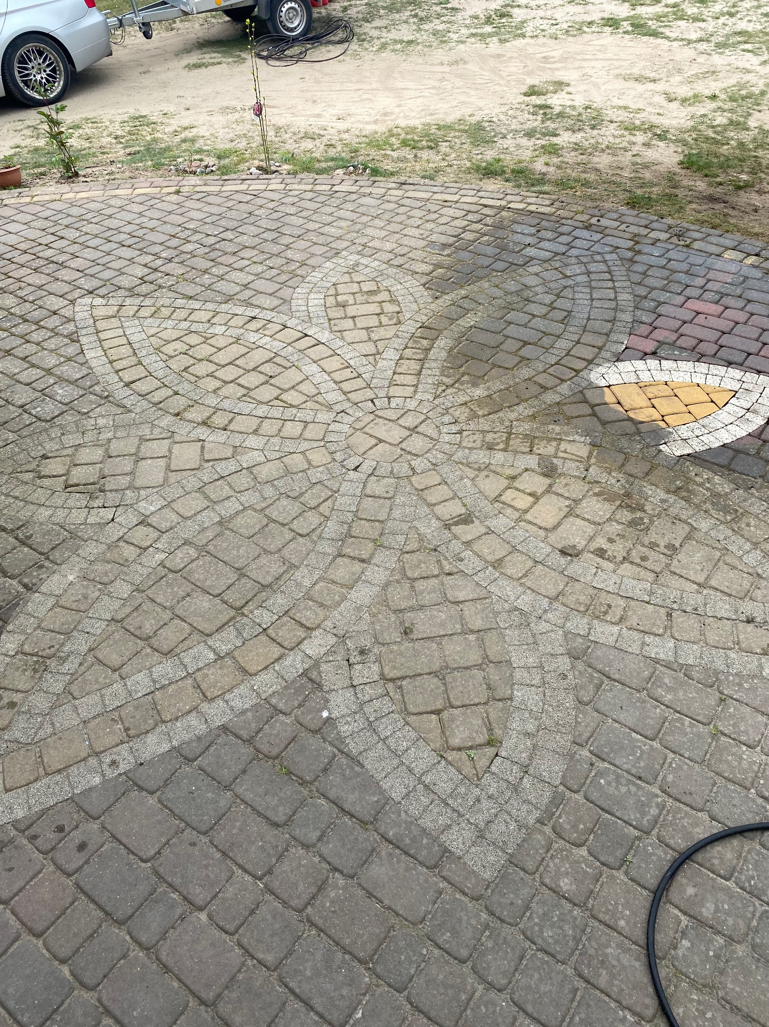 Czyszczona kostka brukowa z wzorem kwiatowym. Część kostki jest mokra, ukazując efekt czyszczenia. Widoczny wąż i fragment samochodu.