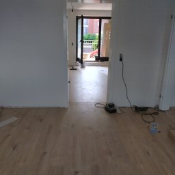 Ad-bud - Jasne panele podłogowe w nowym wnętrzu, widok na balkon przez otwarte drzwi. Świeżo pomalowane ściany i narzędzia sugerują prace wykończeniowe.