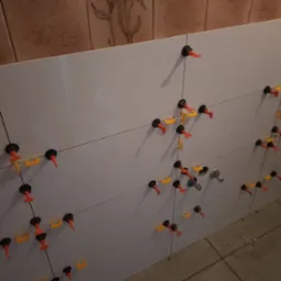 Białe płytki ścienne w trakcie montażu, z użyciem systemów poziomowania płytek. Widoczne fugi i fragment instalacji wodnej.