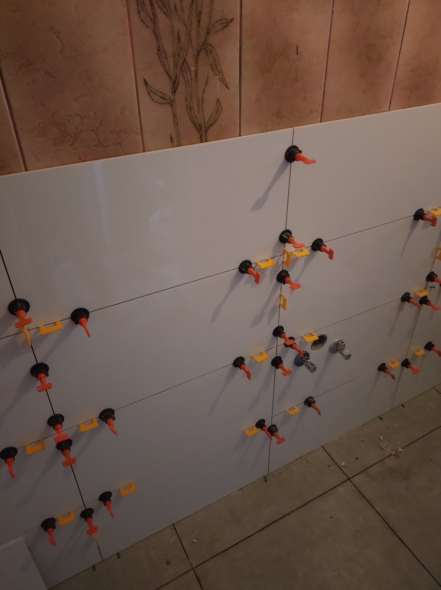 Białe płytki ścienne w trakcie montażu, z użyciem systemów poziomowania płytek. Widoczne fugi i fragment instalacji wodnej.