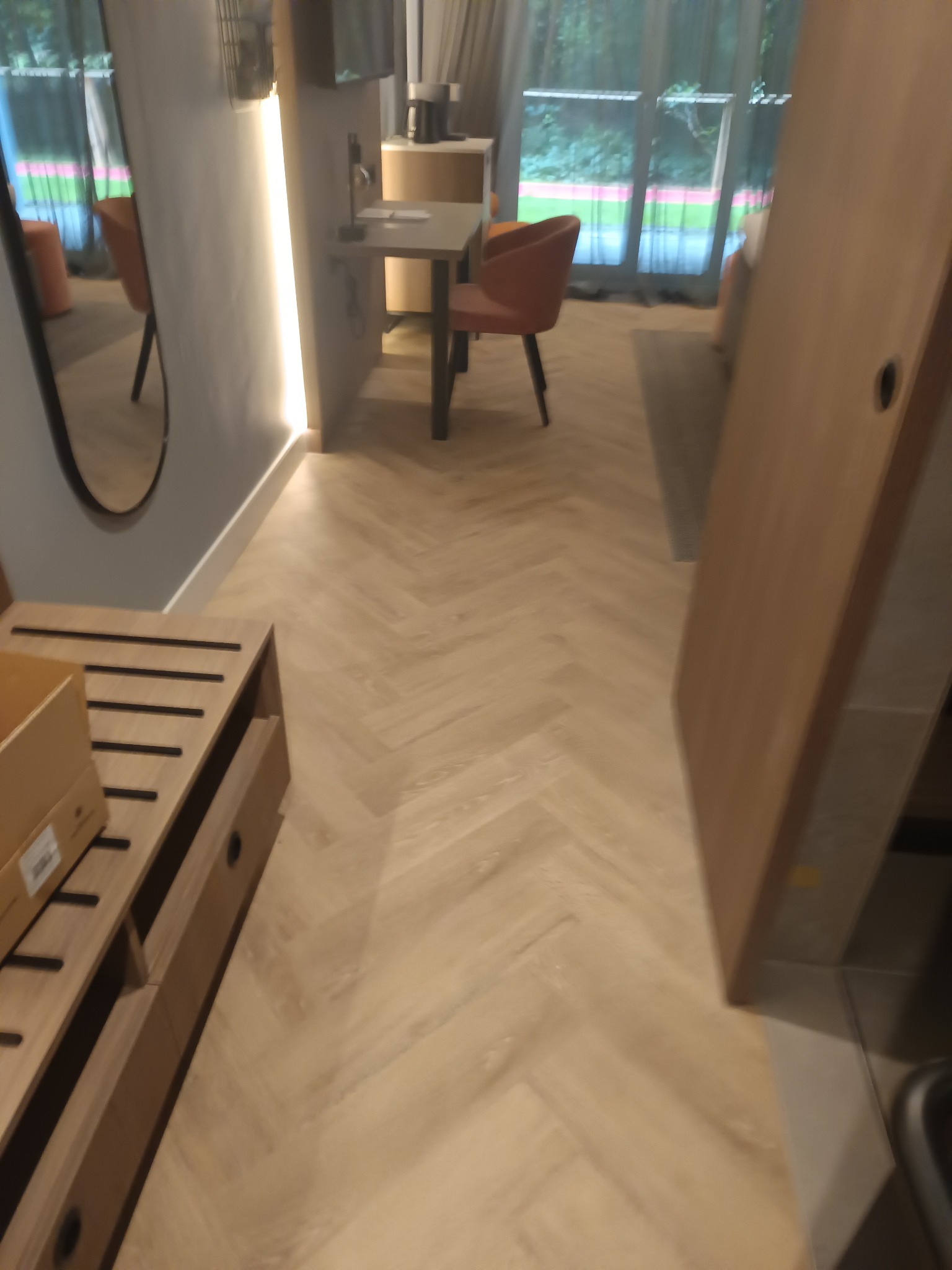 Jasny parkiet ułożony w jodełkę francuską w nowoczesnym wnętrzu. Widoczny fragment mebla z szufladami i krzesło przy stole. W tle okno z widokiem na zieleń.