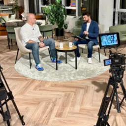 Dwóch mężczyzn prowadzi rozmowę podczas sesji nagraniowej z użyciem profesjonalnego sprzętu filmowego i monitora podglądowego w nowoczesnym wnętrzu.