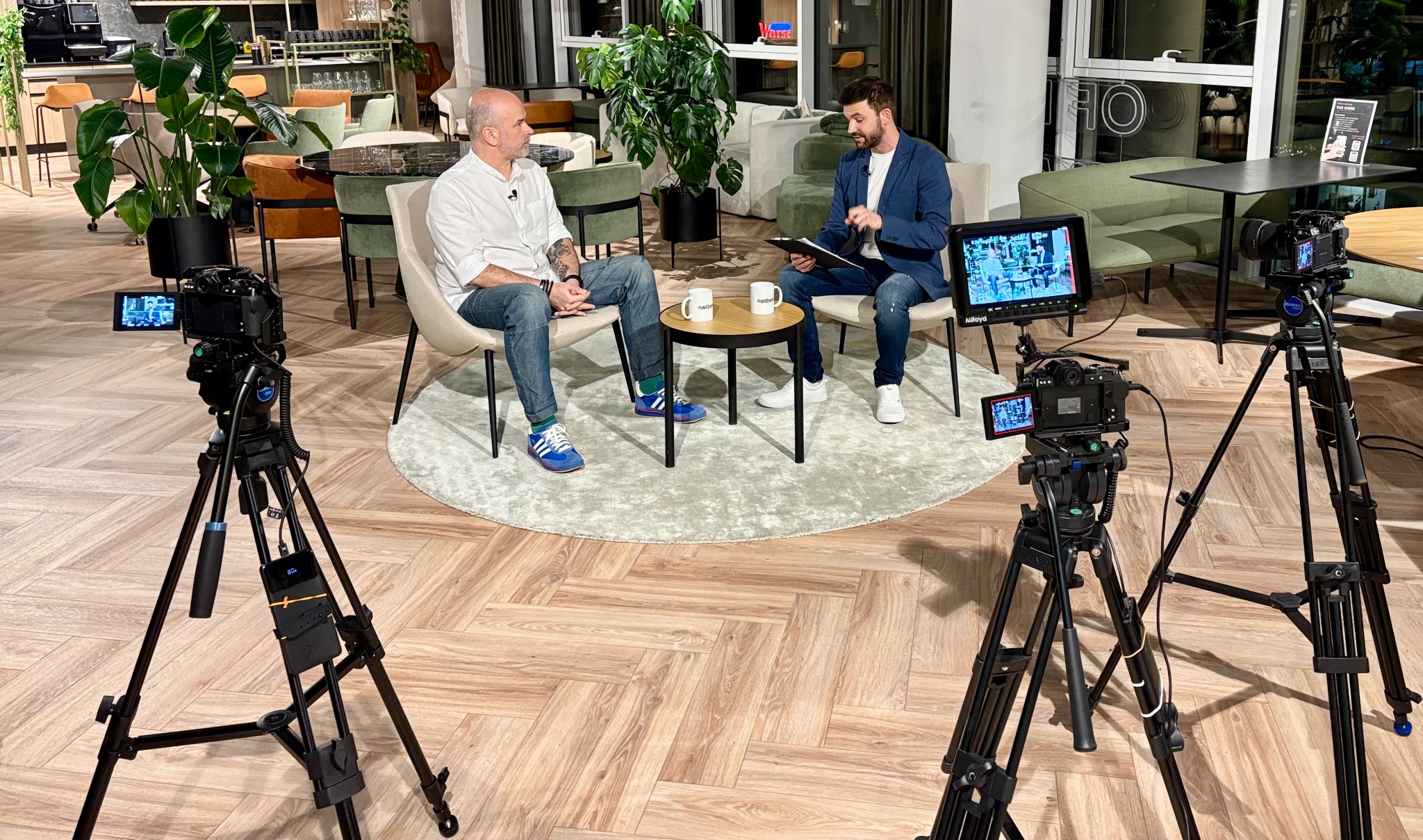 Dwóch mężczyzn prowadzi rozmowę podczas sesji nagraniowej z użyciem profesjonalnego sprzętu filmowego i monitora podglądowego w nowoczesnym wnętrzu.