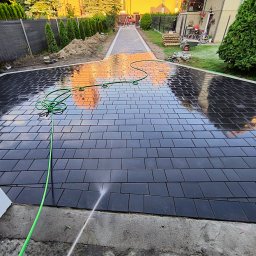 BrukON - Mycie czarnej kostki brukowej na podjeździe domu. W tle widoczny ogród z tujami i ogrodzenie panelowe. Woda spływa po powierzchni kostki.