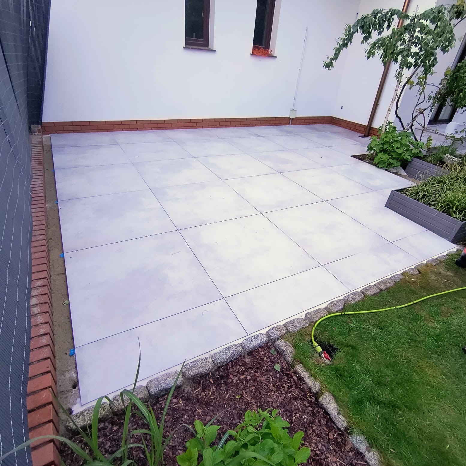 Jasnoszara, ułożona płytka na tarasie przydomowym, obrzeża z kostki granitowej, widoczna zieleń i fragment ściany budynku z oknami.