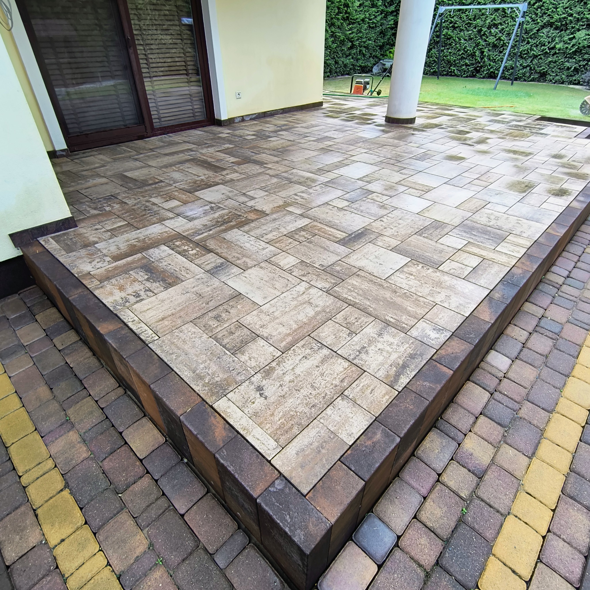 Ukończony taras z jasnobrązowej kostki brukowej ułożonej we wzór jodełki, z ciemnobrązowym obramowaniem. Widoczny fragment elewacji domu i trawnik w tle.