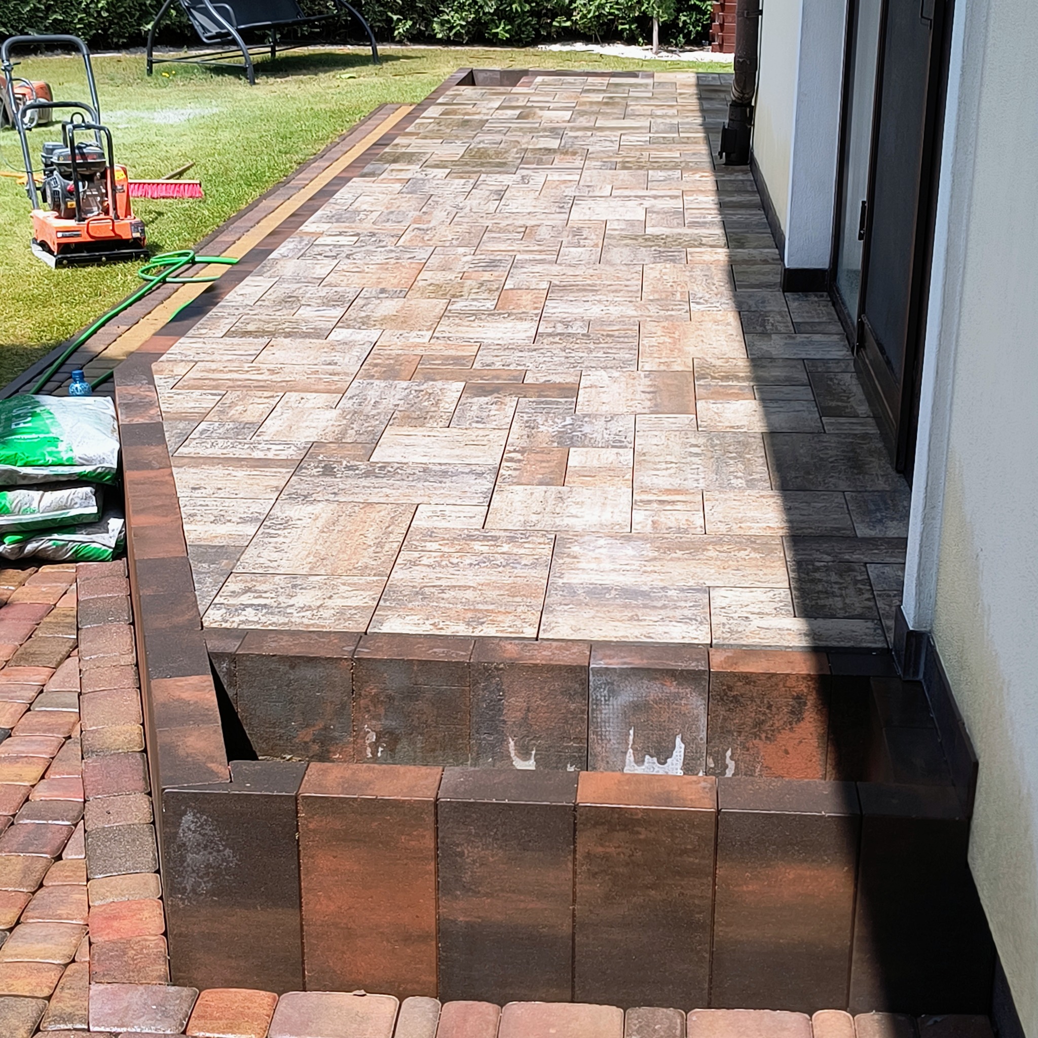 Ułożona kostka brukowa w odcieniach brązu i beżu tworzy ścieżkę przy budynku. Na trawniku stoi zagęszczarka i leżą worki z materiałem.