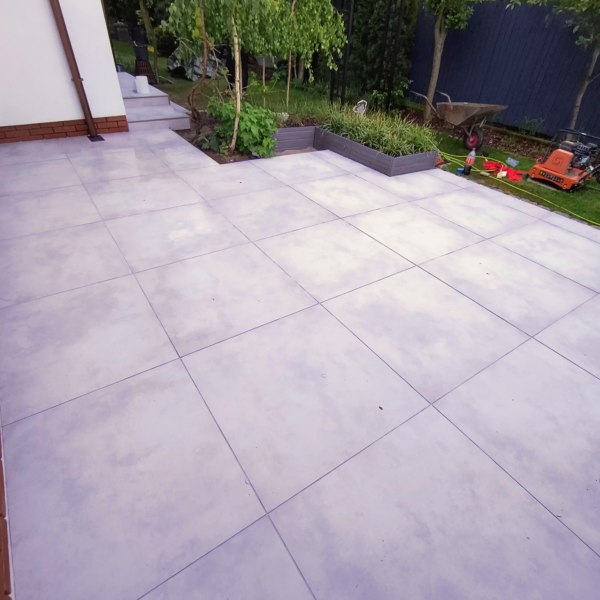 Wykończony taras z jasnoszarej, dużej płytki, z elementami zieleni w donicach i sprzętem ogrodniczym w tle. Widok na ogród i fragment domu.