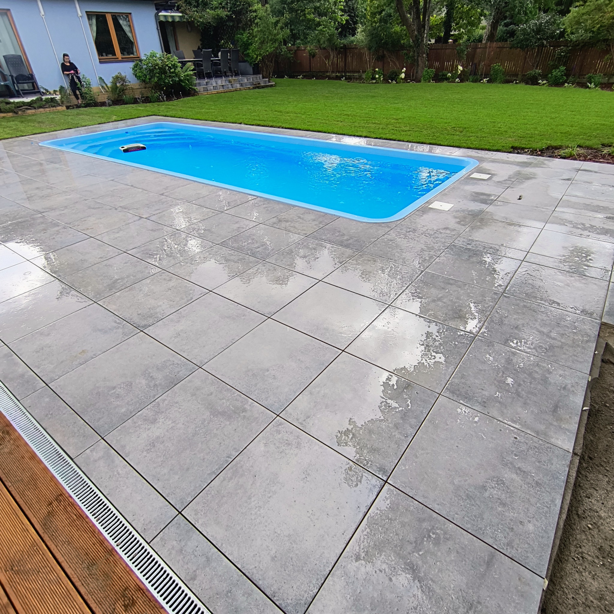 Nowoczesny basen otoczony szarą kostką brukową, widok z góry. W tle zadbany trawnik i zieleń. Fragment drewnianego tarasu z odpływem liniowym. Osoba w tle.