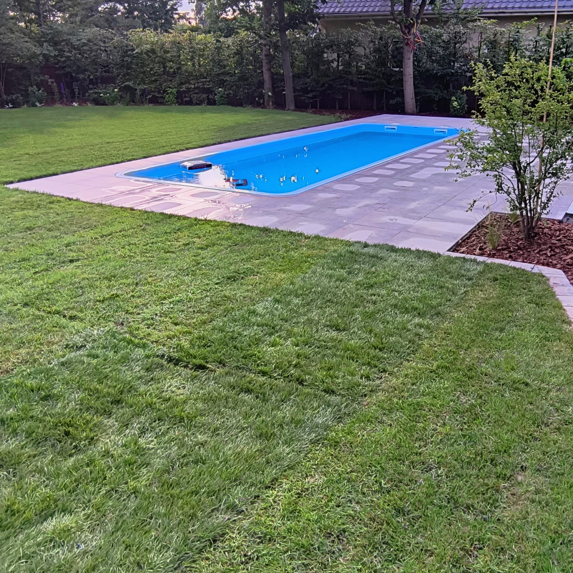 Narożny basen z błękitną wodą otoczony kostką brukową i trawnikiem, widok z poziomu trawy. Elementy małej architektury ogrodowej.