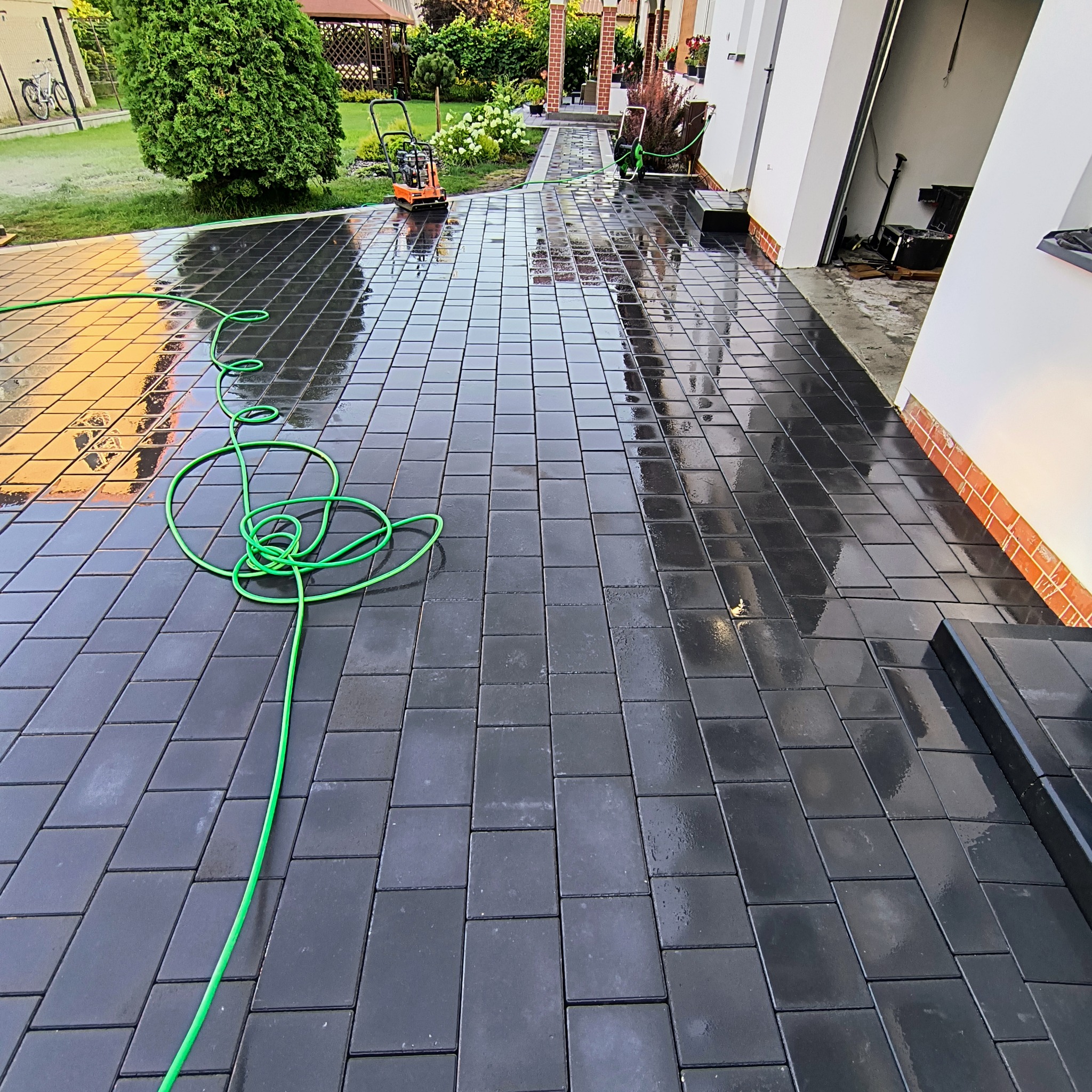 Świeżo ułożona, czarna kostka brukowa odbija światło, z zielonym wężem ogrodowym w pierwszym planie. W tle widoczna ubijarka wibracyjna i budynek.
