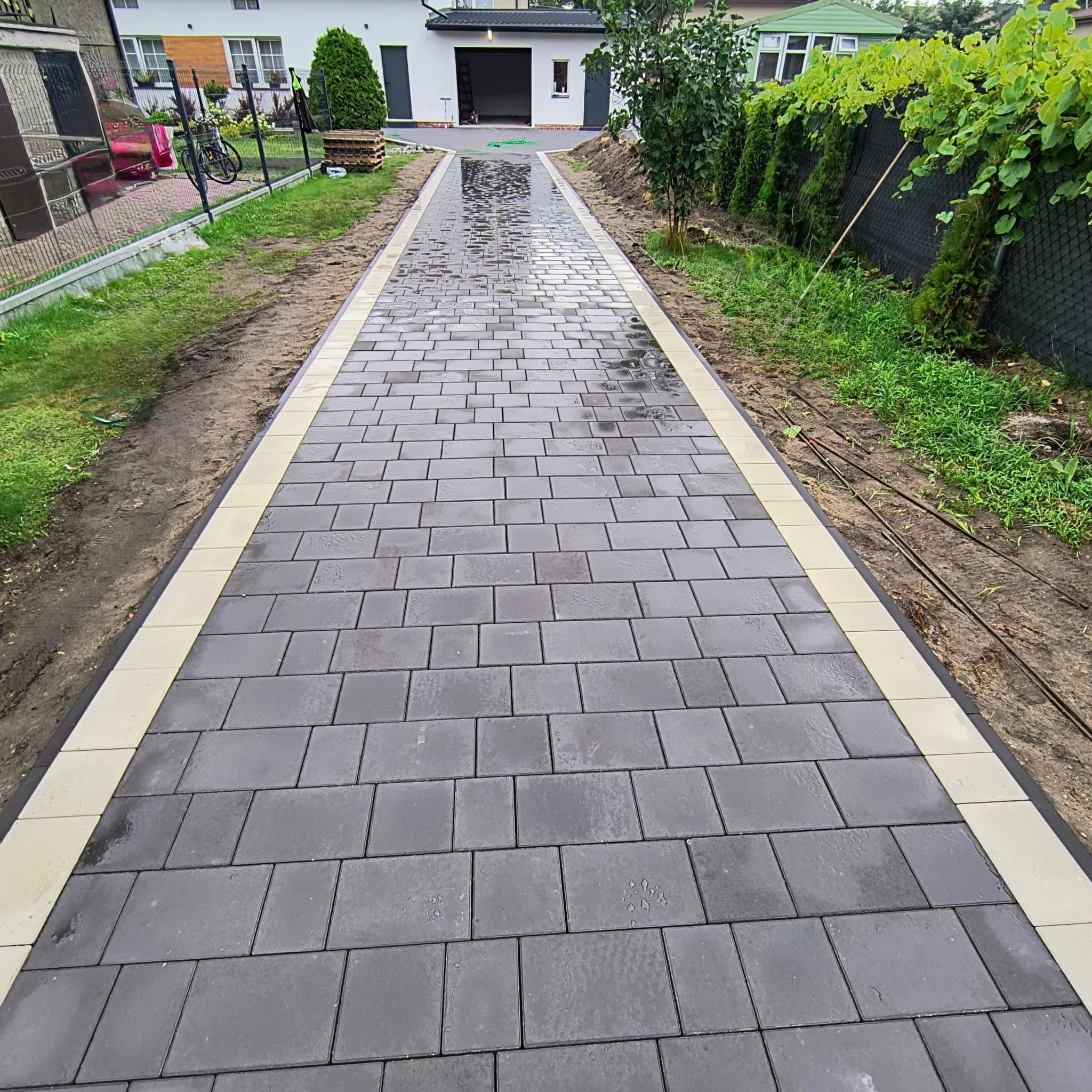 Nowo ułożona, mokra, szara kostka brukowa z jasnymi obrzeżami prowadząca do garażu. Widoczne trawniki po bokach i fragment ogrodzenia z roślinami.