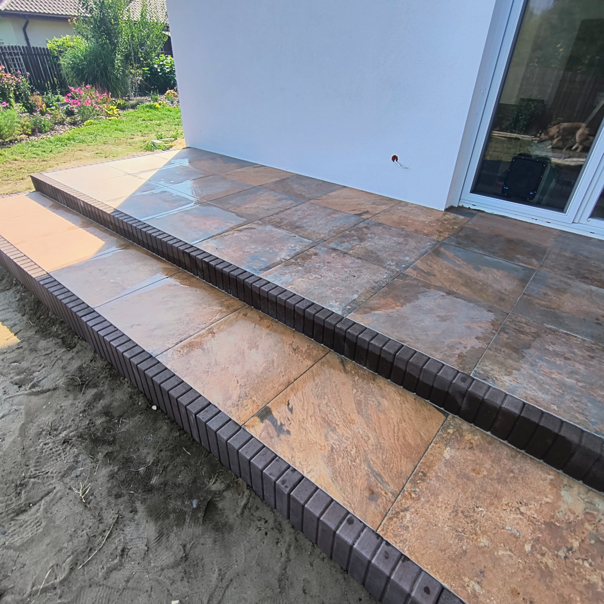 Wykończone schody z płytek imitujących kamień, z ciemnobrązową obwódką z kostki, prowadzące do drzwi tarasowych. Widoczny fragment ogrodu i elewacji domu.