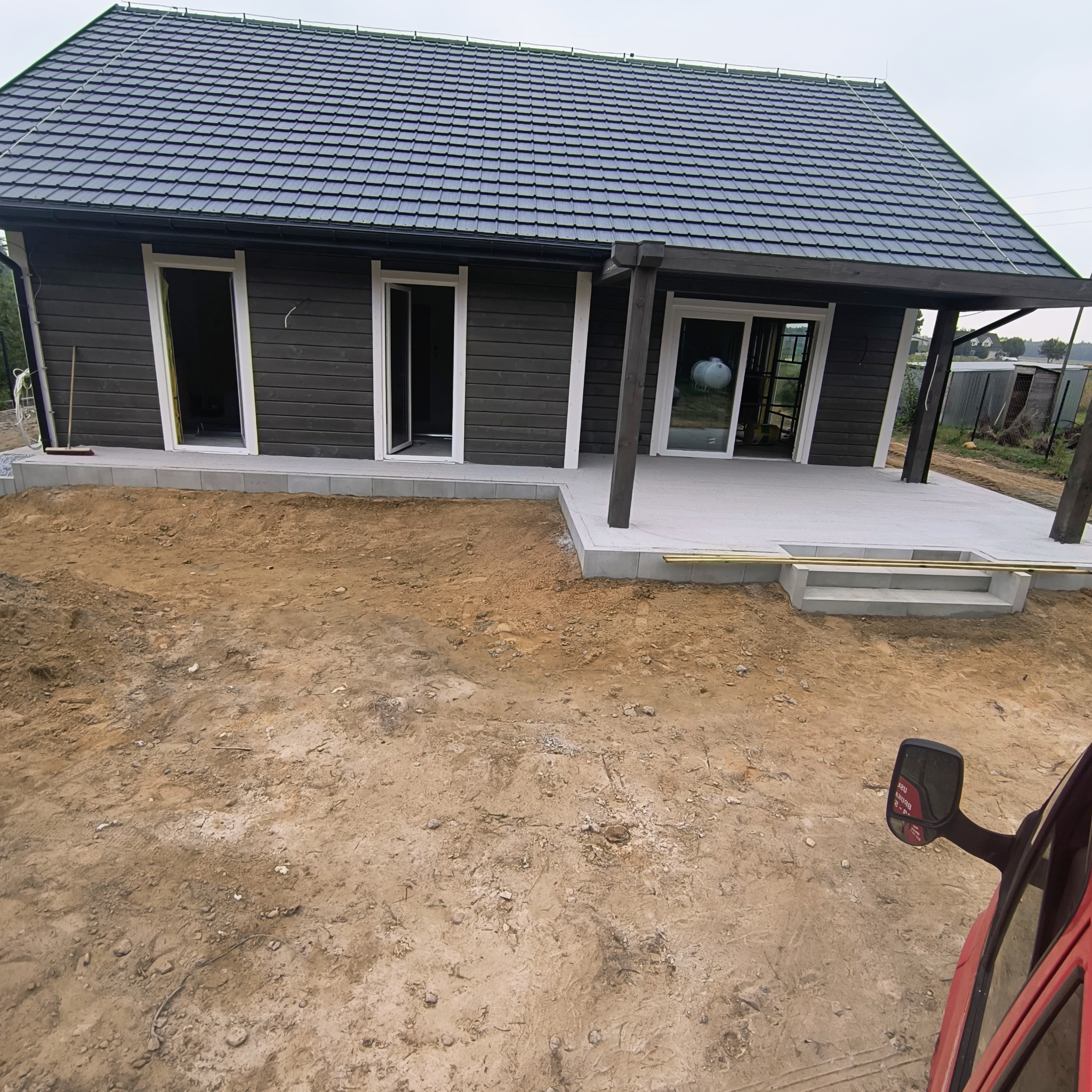 Dom z ciemnego drewna z betonowym tarasem w trakcie budowy. Widoczne schody i przygotowany teren pod ogród.