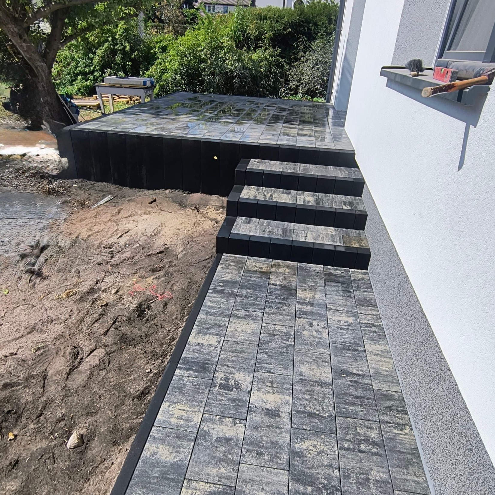 Ułożona kostka brukowa w odcieniach szarości tworząca ścieżkę i schody prowadzące do podwyższonego tarasu. Widoczny fragment elewacji budynku i teren przygotowany pod nasadzenia.