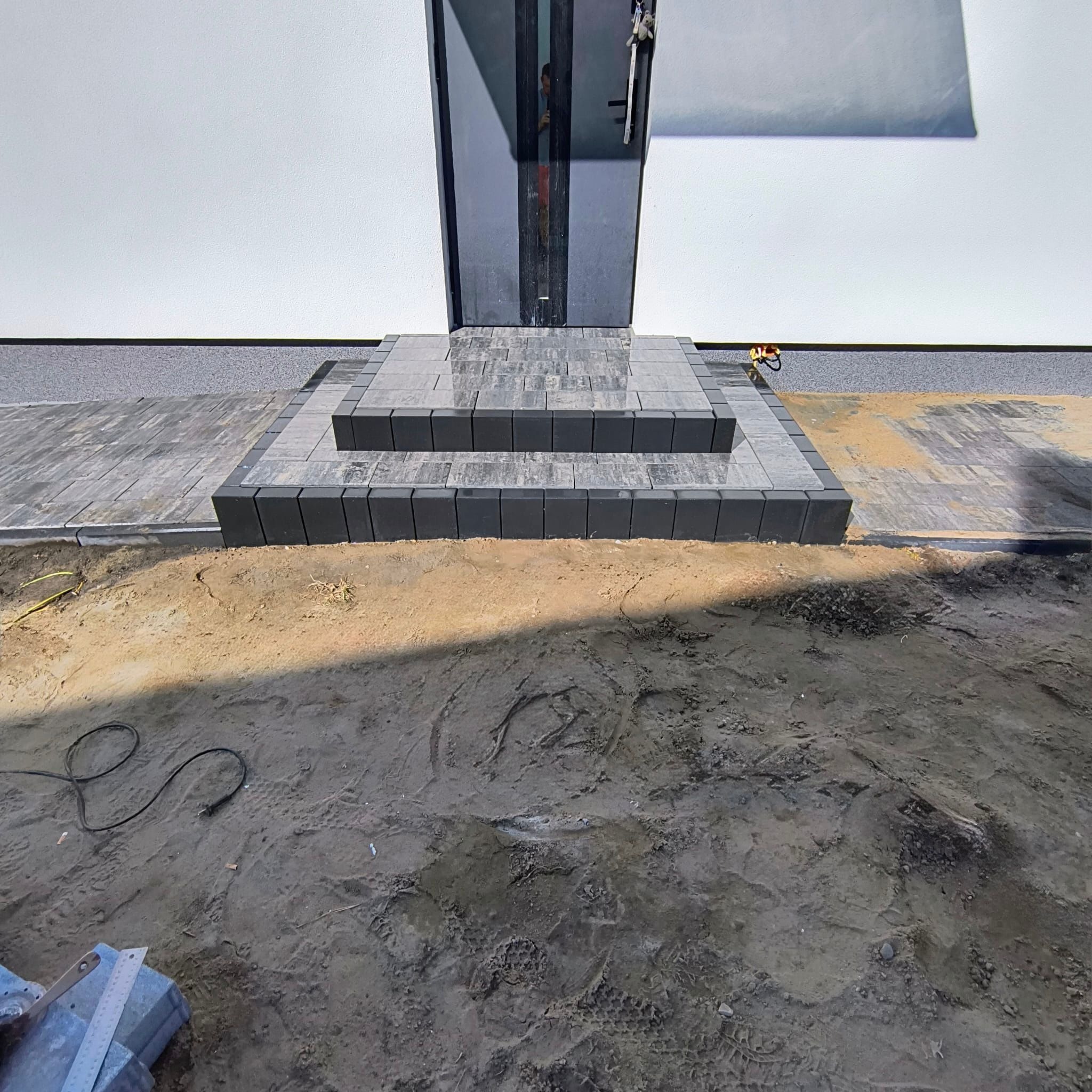 Schody z kostki brukowej prowadzące do drzwi wejściowych, widoczne ślady prac ziemnych. Kostka w odcieniach szarości, świeżo ułożona, zarys budynku w tle.