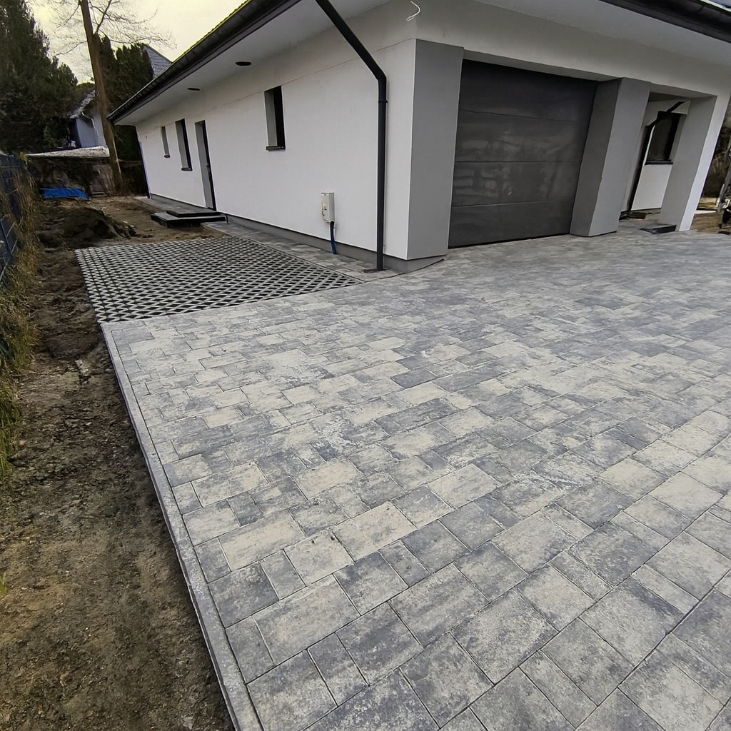 Ułożona szara kostka brukowa przed nowoczesnym domem z garażem. Fragment czarnej kostki ażurowej przy ścianie budynku. Widok z boku, ziemia po lewej stronie.