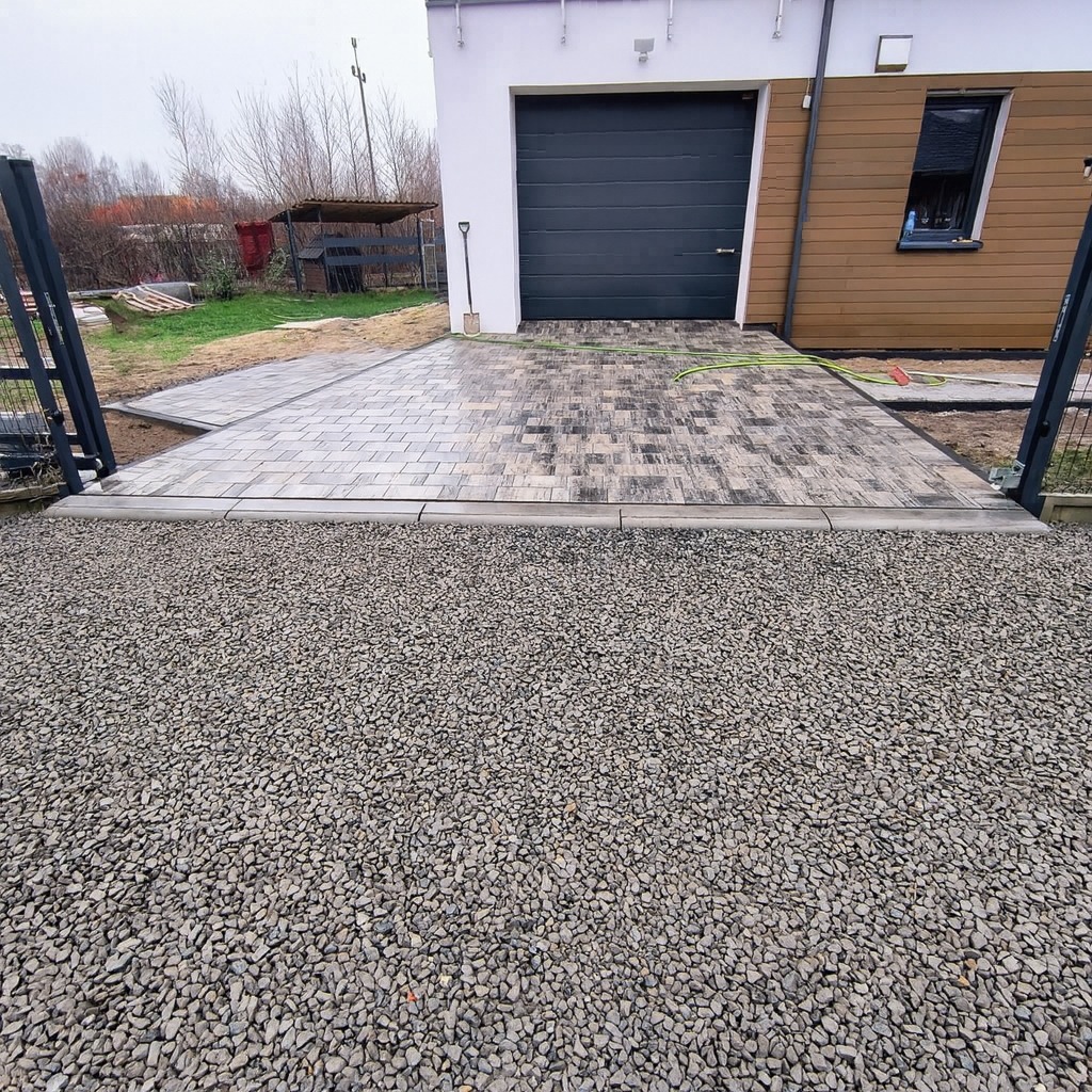 Ułożona kostka brukowa na podjeździe do garażu, z widocznym fragmentem ogrodzenia i wysypanym żwirem. Kostka w odcieniach szarości i brązu, ułożona we wzór.