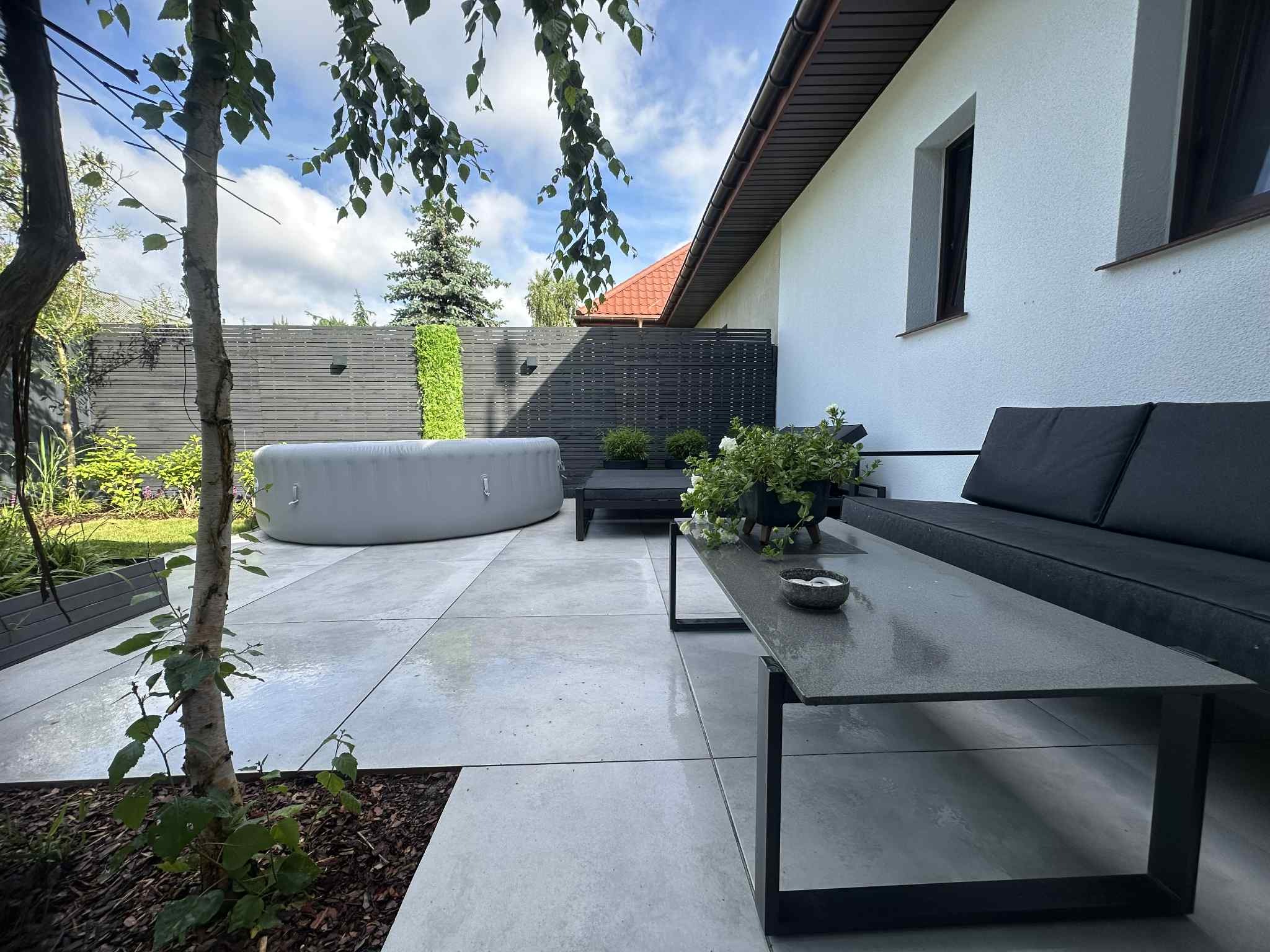 Nowoczesny taras z szarych płyt, otoczony zielenią i drewnianym ogrodzeniem. W tle widoczna wanna SPA. Minimalistyczny design z elementami roślinnymi.