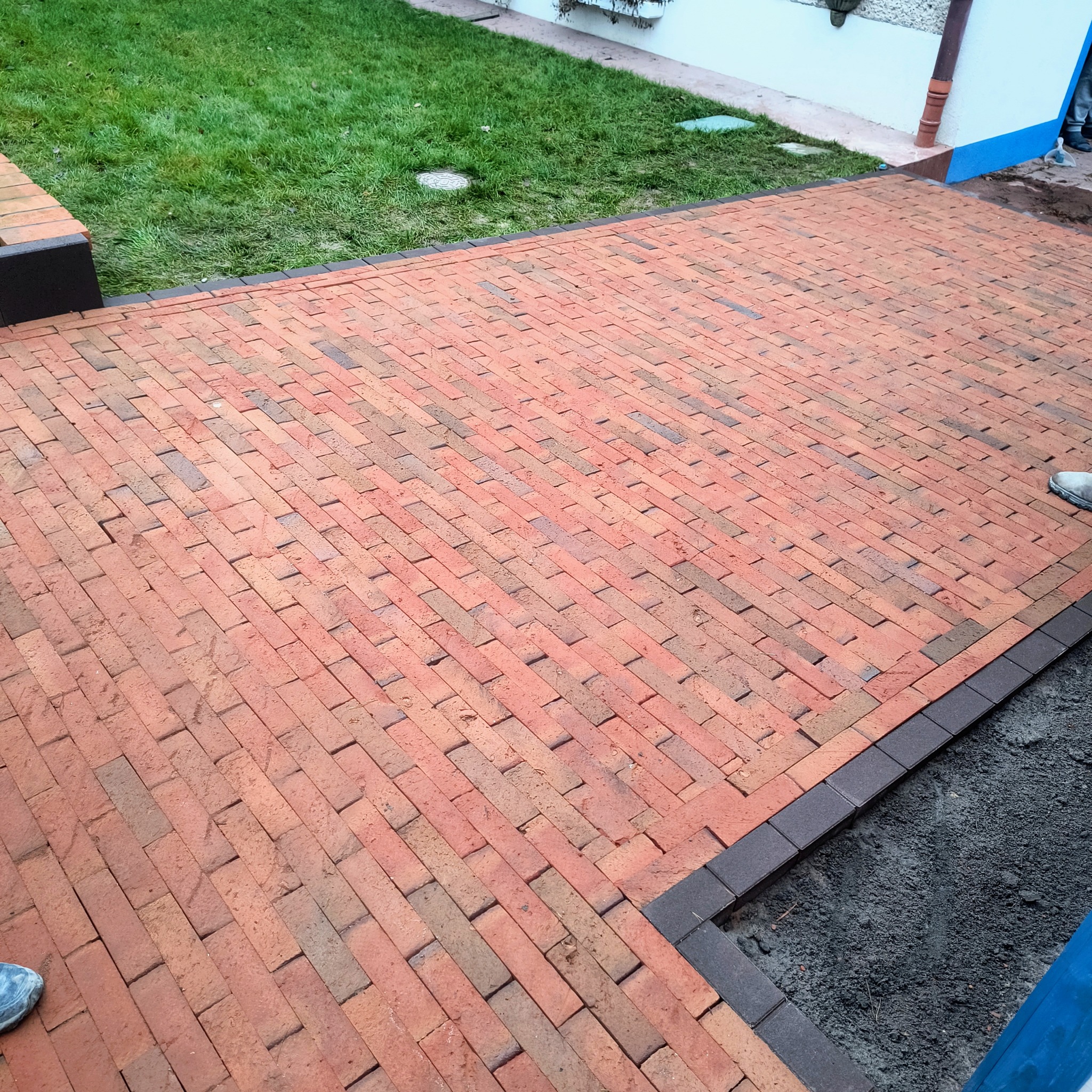 Ukończona nawierzchnia z czerwonej kostki brukowej ułożonej w jodełkę, obrzeża z ciemnej kostki. Widok na trawnik i fragment budynku w tle.