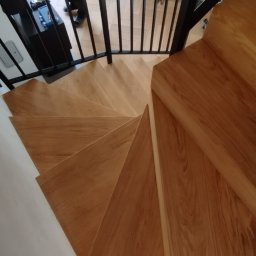 Schody drewniane YEVHENII ALIEKSIEIEV - Kątowe ujęcie drewnianych schodów spiralnych z czarną metalową balustradą. Widoczne detale wykończenia i naturalne usłojenie drewna. W tle osoba i fragment salonu.