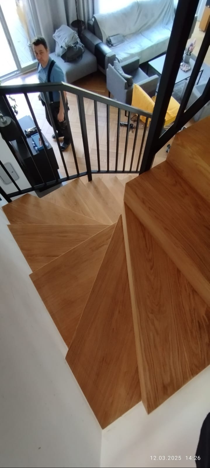 Kątowe ujęcie drewnianych schodów spiralnych z czarną metalową balustradą. Widoczne detale wykończenia i naturalne usłojenie drewna. W tle osoba i fragment salonu.