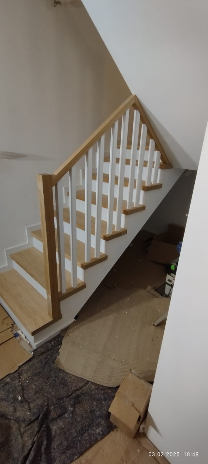 Jasne schody z drewnianymi stopniami i białą balustradą w nowym domu, widok z dołu. Widoczne zabezpieczenia podłogi na czas budowy.