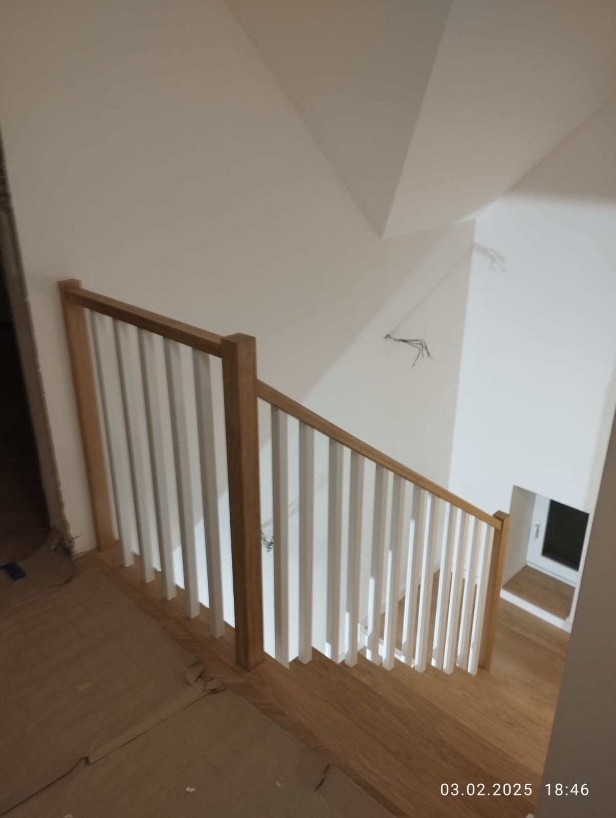 Nowoczesne schody z jasnego drewna z białą balustradą, widok z góry. Fragment schodów zabezpieczony kartonem. W tle widoczne okno.