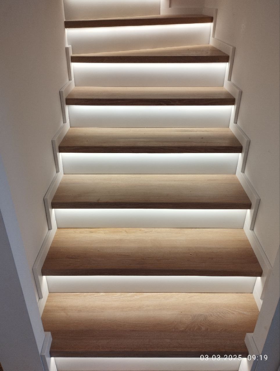 Nowoczesne schody z drewna z subtelnym podświetleniem LED. Jasne drewno i białe elementy tworzą minimalistyczny, elegancki design. Widok z dołu na stopnie.