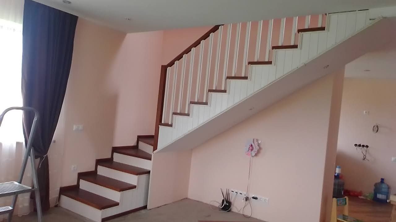 Jasne schody z drewnianymi stopniami i białą balustradą w jasnym wnętrzu. Widoczne gniazdka elektryczne i dekoracja ścienna w kształcie kwiatka.