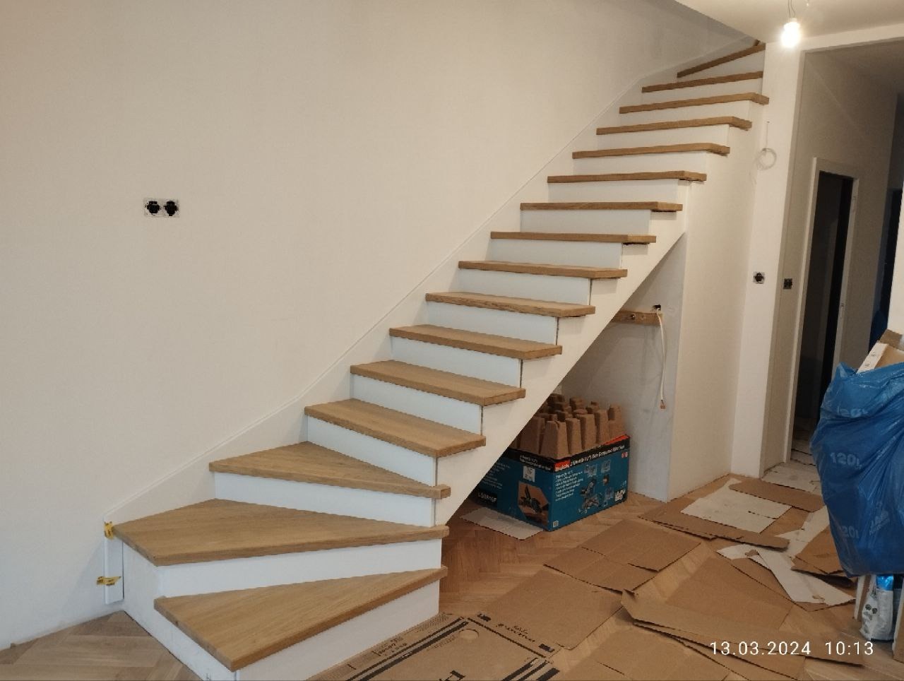 Nowoczesne schody wewnętrzne z drewnianymi stopniami i białymi podstopnicami, zamontowane w minimalistycznym wnętrzu. Widoczne przygotowania do dalszych prac wykończeniowych.