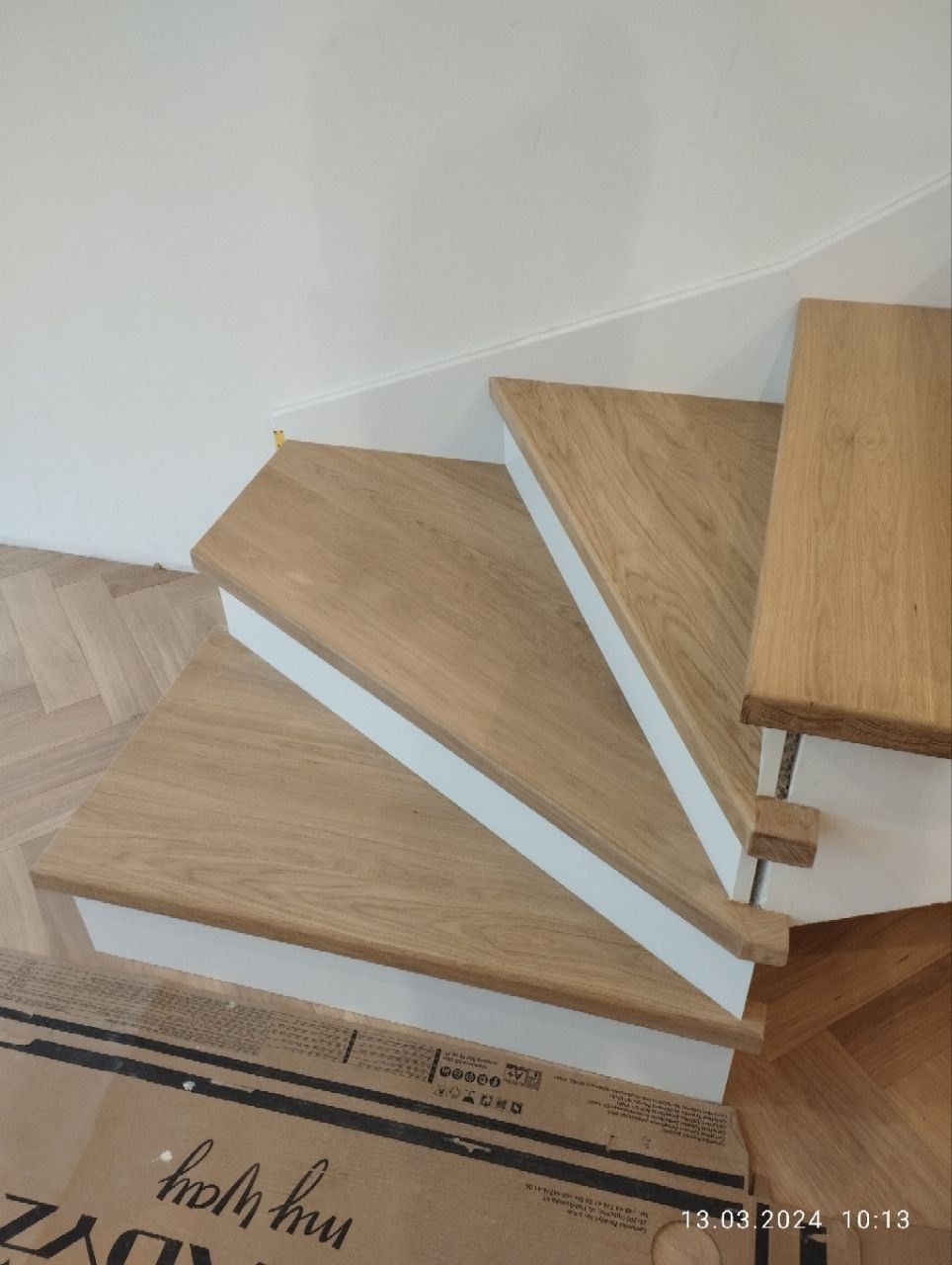 Jasne, drewniane schody z białymi podstopnicami w nowoczesnym domu, widoczne z góry, z fragmentem parkietu i kartonem w tle.