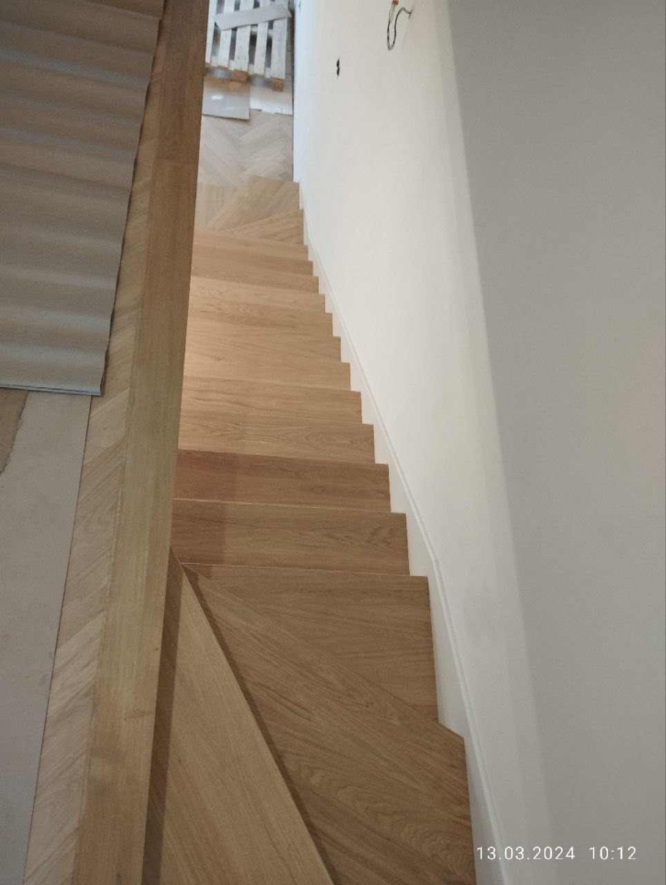 Jasne, dębowe schody bez balustrady, widok z góry. Stopnie z wyraźnym usłojeniem. Podłoga parkiet w jodełkę. Białe ściany i listwy przypodłogowe. Surowy przewód elektryczny.