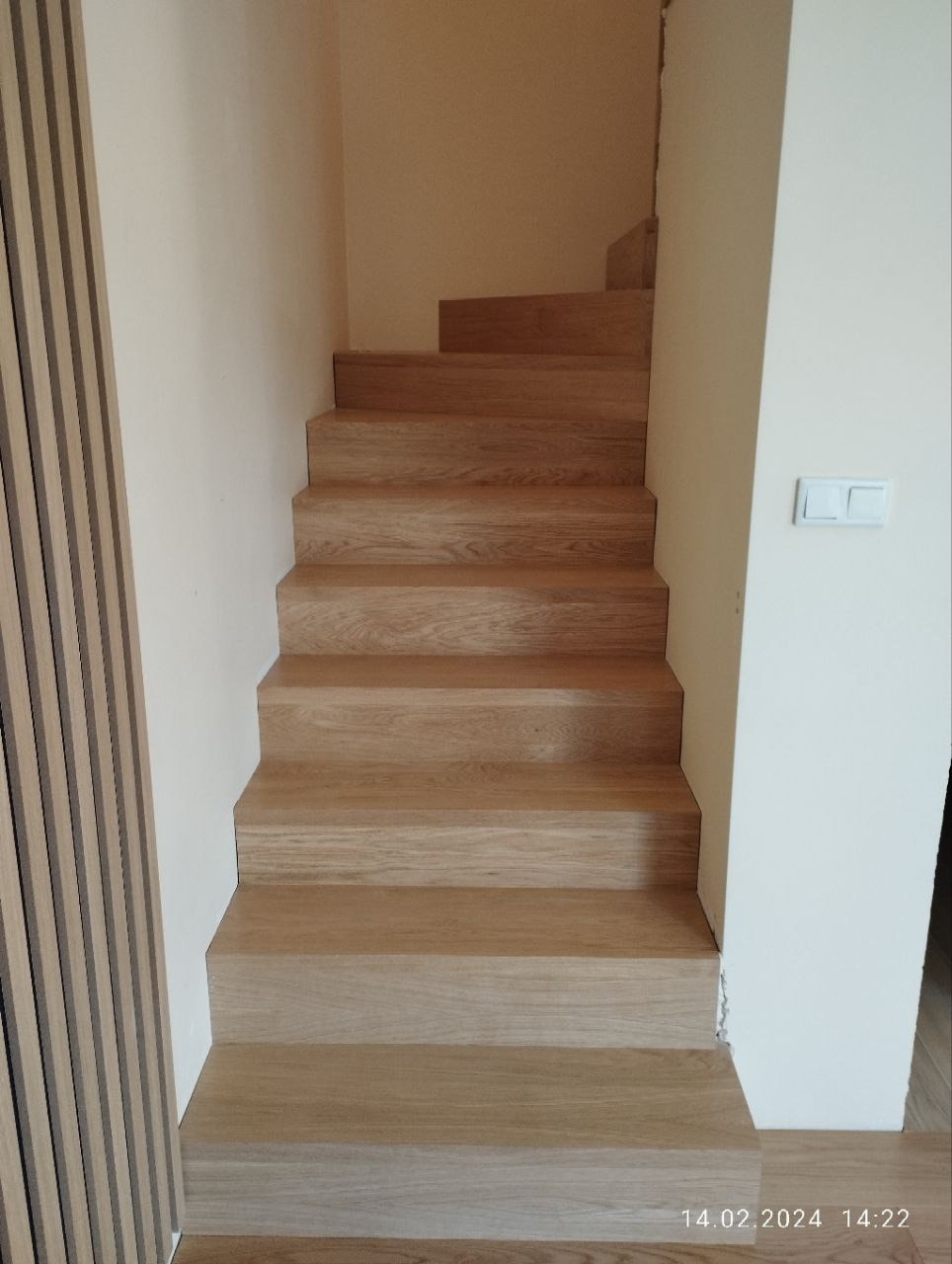 Jasne drewniane schody bez balustrady, prowadzące do góry, z widocznym fragmentem dekoracyjnej okładziny ściennej po lewej stronie i białym włącznikiem światła po prawej.