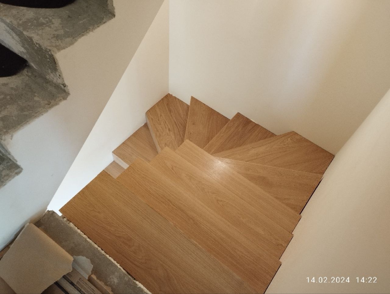 Widok z góry na jasne, drewniane schody w trakcie montażu, z widocznym surowym betonem i fragmentem ściany. Oświetlenie naturalne, ciepłe barwy drewna.