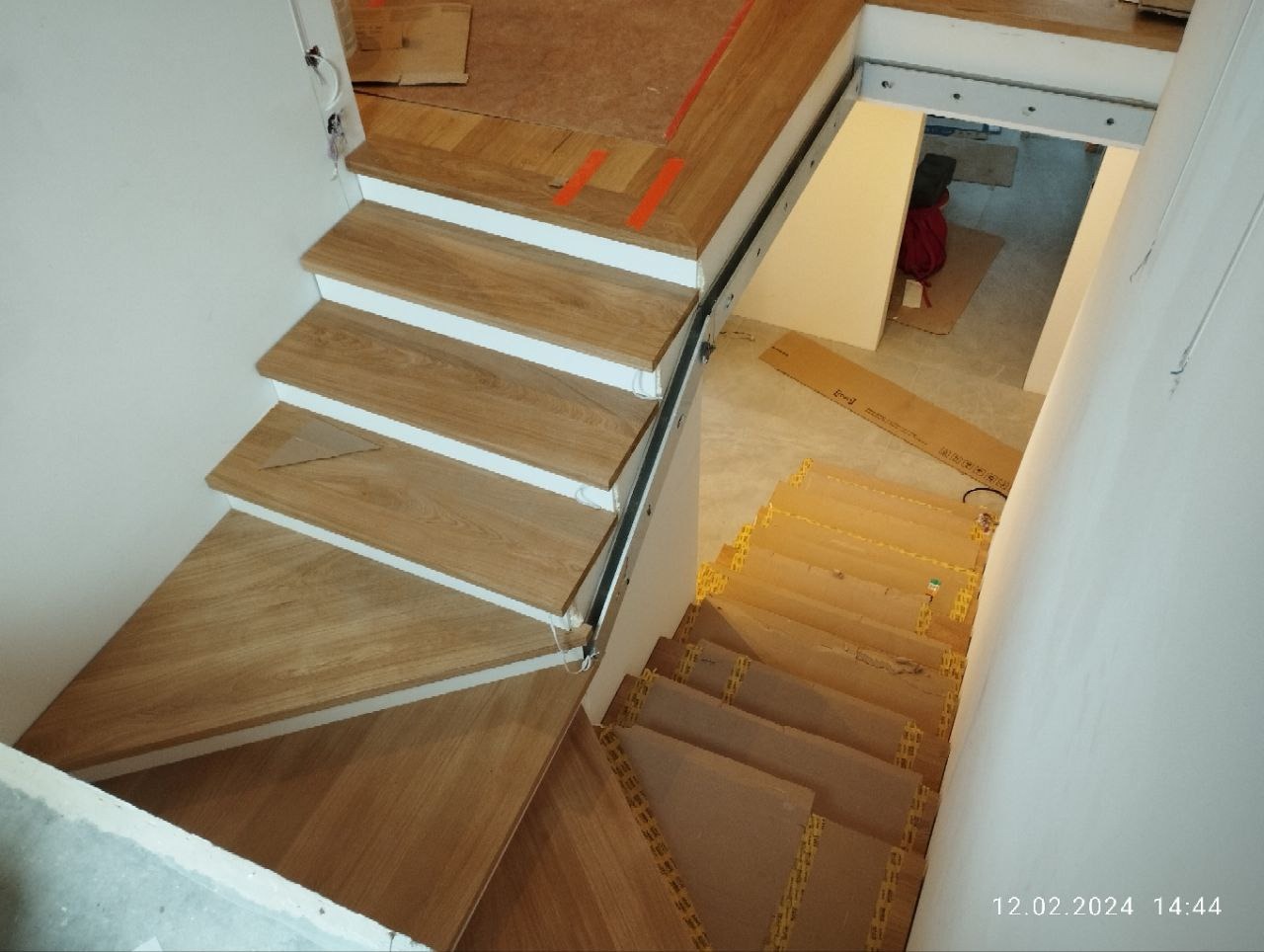 Jasne, drewniane schody z białą obudową i czarną poręczą, widok z góry na dół, z widocznym fragmentem piwnicy w tle. Minimalistyczny design.