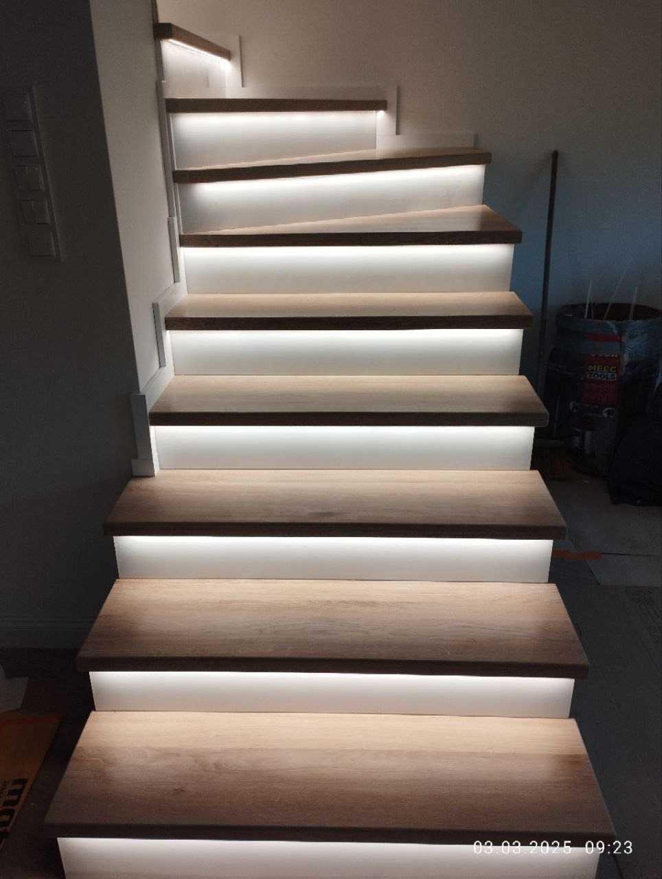 Nowoczesne schody z drewna z subtelnym podświetleniem LED, widok od dołu. Stopnie w ciepłym odcieniu drewna kontrastują z białymi podstopnicami.