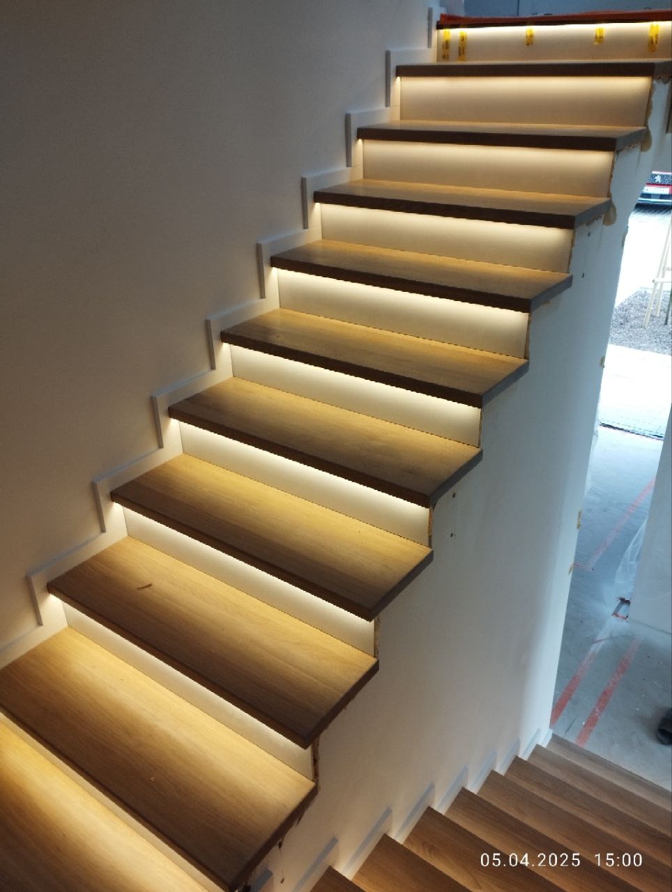 Nowoczesne schody z drewna z subtelnym oświetleniem LED pod każdym stopniem, widok z dołu, ukazujący detale wykończenia i ciepłą barwę światła.