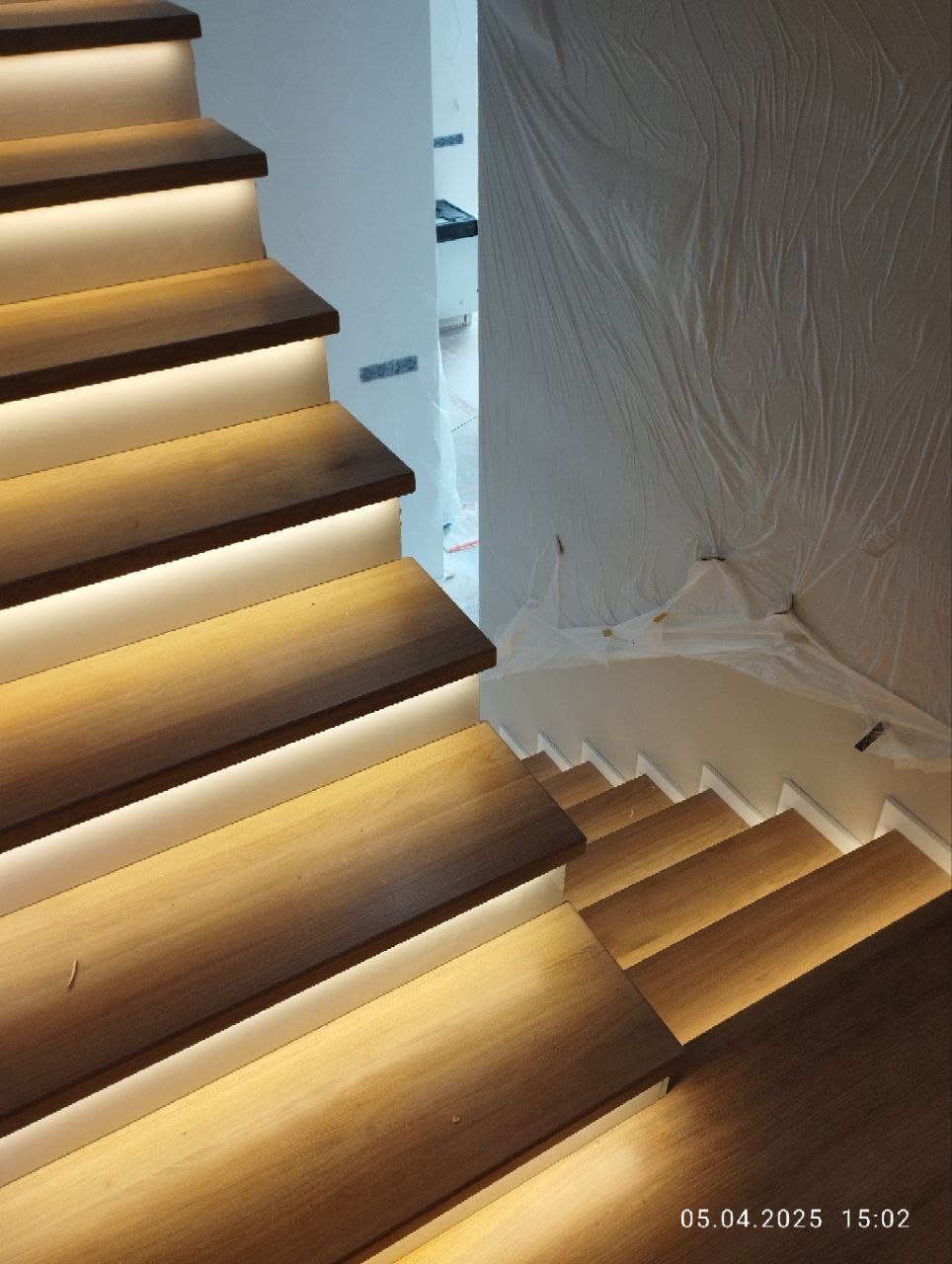 Nowoczesne schody z drewna z podświetleniem LED, widok z góry. Ściany zabezpieczone folią malarską, sugerujące świeżo zakończony montaż.