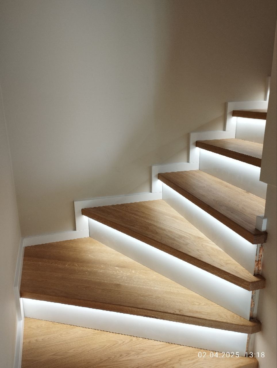 Nowoczesne schody z drewna dębowego z białymi podstopnicami i subtelnym oświetleniem LED, tworzącym elegancki efekt wizualny. Ujęcie z dołu, akcentujące detale wykończenia.