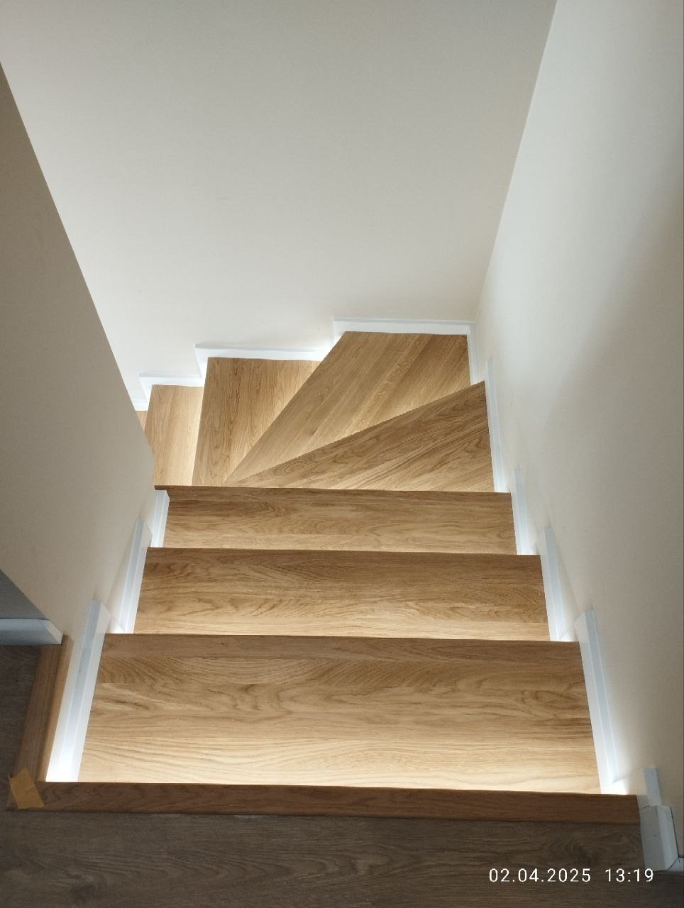 Nowoczesne schody z jasnego drewna, podświetlane białym światłem LED, widok z góry. Minimalistyczny design, neutralna kolorystyka ścian i sufitu.