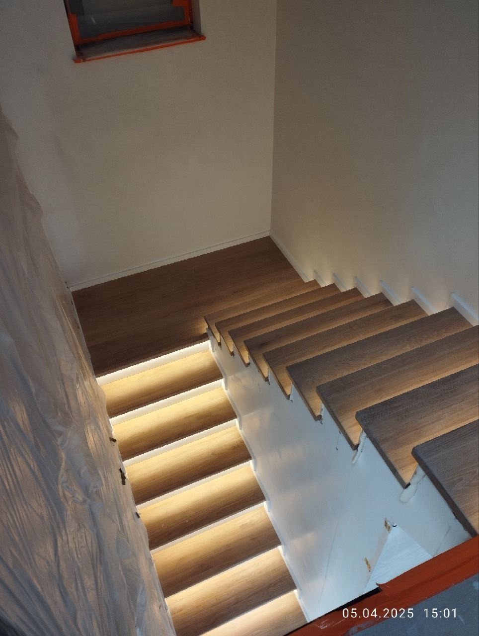 Nowoczesne schody z podświetleniem LED, widok z góry. Część ściany zabezpieczona folią malarską. Jasne drewno i białe wykończenie.